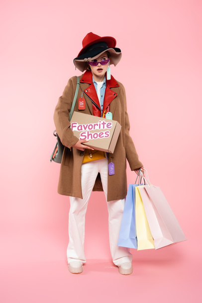 surprised woman in sunglasses and hats with sale tags holding box with favorite shoes lettering and shopping bags on pink, black friday concept - Foto, afbeelding