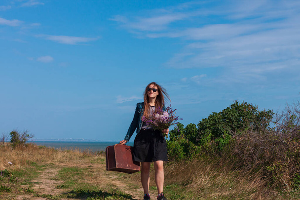 Young woman with long brown hair in denim jacket, black skirt, vintage suitcase,purple wild flowers bouquet off-road sea landscape. Hitchhiker on countryside trip. Lifestyle photo lady walking outdoor - Photo, Image