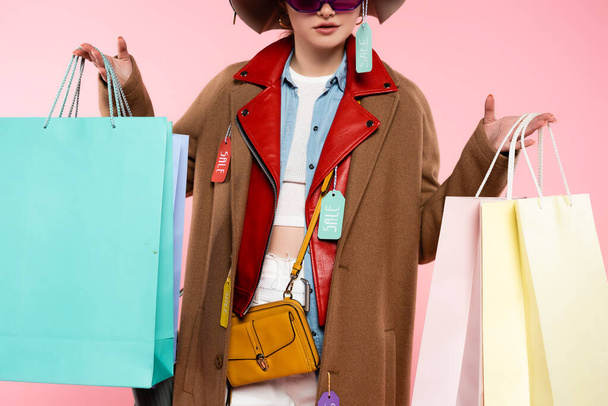 cropped view of trendy woman in sunglasses and hats with sale tags holding shopping bags on pink - Fotografie, Obrázek
