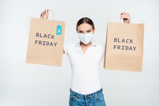customer in medical mask holding shopping bags with sale tag and black friday lettering isolated on white  - Φωτογραφία, εικόνα