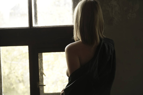 Back view of beautiful lonely   woman in black leather jacket standing near window. - Fotoğraf, Görsel