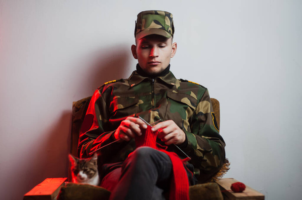 ein junger Kerl in Militäruniform sitzt auf einem Stuhl und strickt in einer ruhigen Umgebung - Foto, Bild
