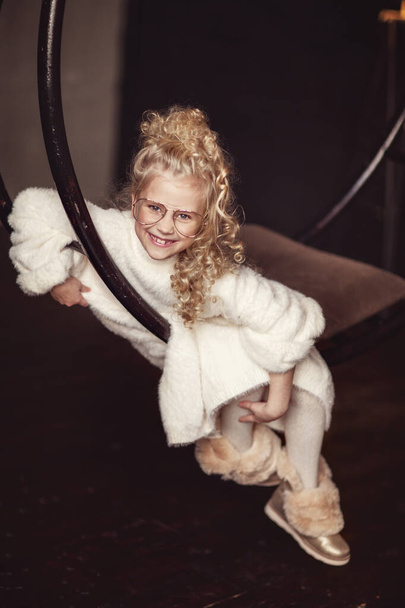 Une belle et drôle blonde bouclée dans un pull moelleux blanc s'assoit sur une balançoire sur un fond sombre, sourit et rit largement. Carte postale festive. Joyeux Noël et bonne année. - Photo, image