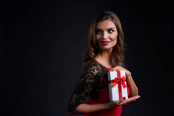 Holidays and celebration concept. Happy gorgeous woman with red lipstick makeup holding gift box with red ribbon, looking to side at blank copy space, over dark background - Photo, image
