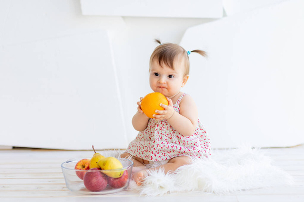 ein kleines Mädchen sitzt in einem hellen Raum mit einem Teller Obst und isst eine Orange - Foto, Bild