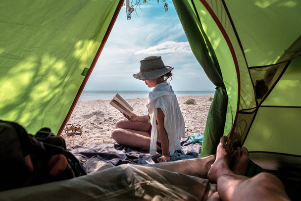 Жінка читає книгу на пляжі біля туристичного намету
 - Фото, зображення