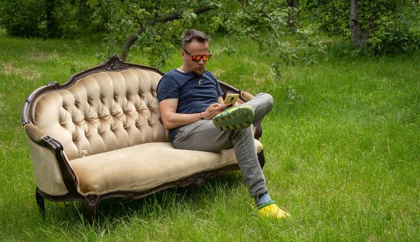 Middle-aged man uses mobile phone resting on light sofa in garden. Sofa is on green grass outdoors. High quality photo - Photo, Image