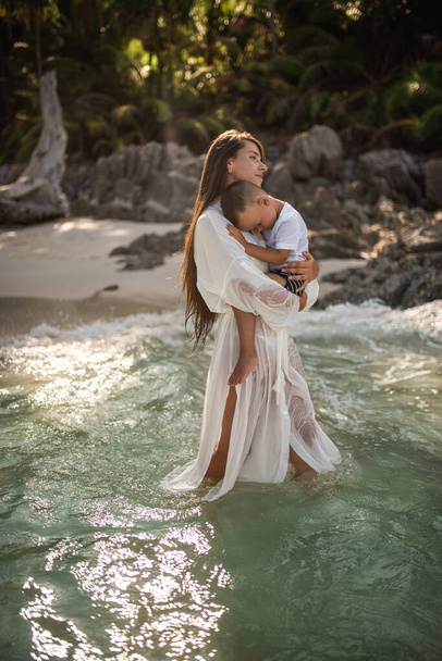 Η μητέρα περπατά κατά μήκος της καταγάλανης θάλασσας και κουβαλάει τον γιο της στην αγκαλιά της - Φωτογραφία, εικόνα