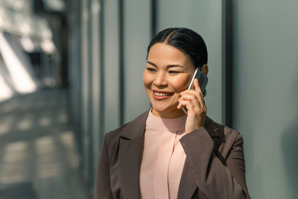 Mulher asiática falando em um telefone celular na rua. Empresária de jaqueta segura telefone na orelha e sorri. Foto de alta qualidade - Foto, Imagem