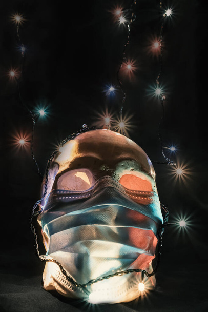 Straszny kompozycja Halloween podczas pandemii koronawirusa. Ludzka czaszka nosząca maskę medyczną na tle kolorowych świateł - Zdjęcie, obraz