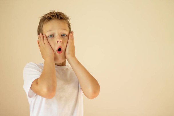 A surprised or scared boy of 7-10 years old in a white T-shirt stands and shouts with his hands on his cheeks on a beige background. Copy space. - Photo, Image
