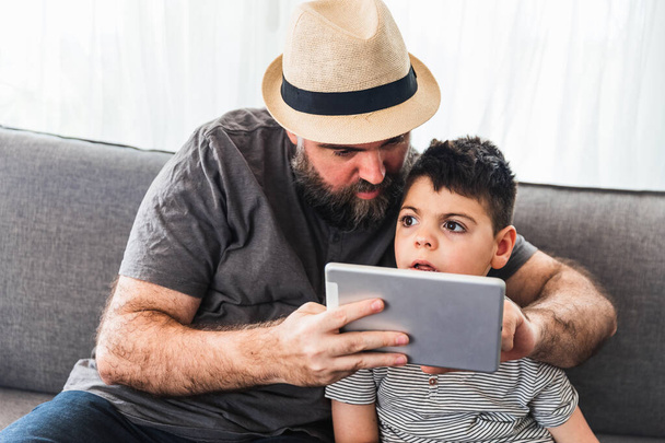 Caucasian bearded man and boy with a tablet on the sofa of a house. Casual dress, with hat. - Photo, Image