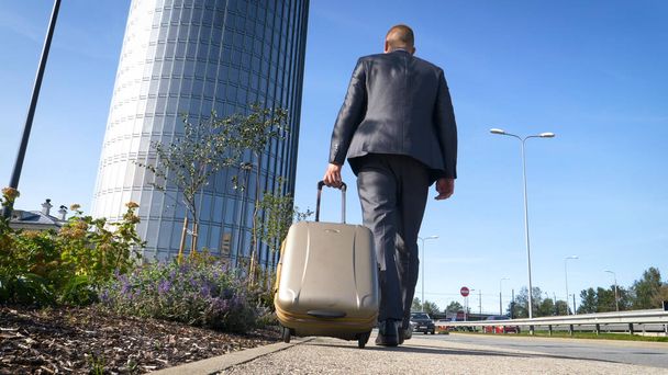Traveling businessman walking with his wheeled suitcase across the city skyline, businessman walking with luggage and using mobile phone. - 写真・画像
