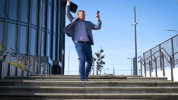 Celebrating success. Low angle view of excited young businessman keeping arms raised and expressing positivity while standing outdoors with office building in the background - Фото, изображение