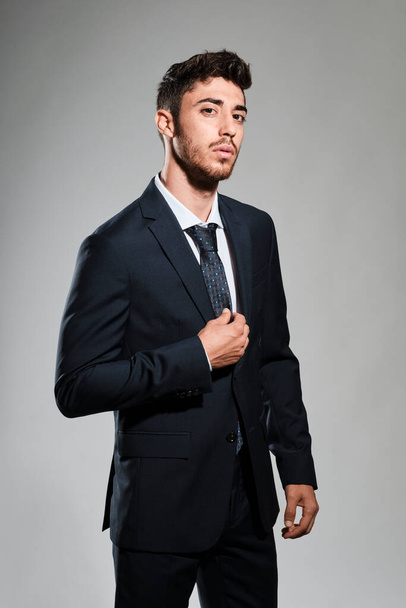 Sakallı, koyu mavi takım elbiseli, sağ eli kravatına dokunan ve kameraya bakan genç bir adam. - Fotoğraf, Görsel