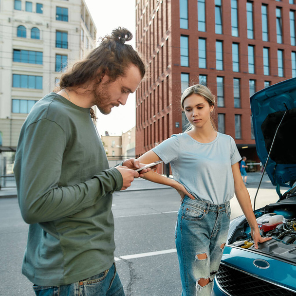 Man decided to help girl with her auto, he is trying to call emergency car service while they both standing near her broken car with open hood on the city street - Foto, Imagem