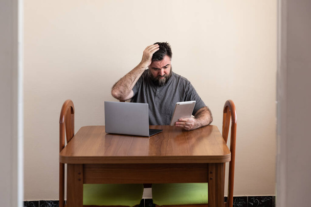 hombre caucásico barbudo sentado frente a una mesa con un ordenador portátil y una tableta. sus brazos están abiertos como signo de duda o duda. camiseta casual - Foto, imagen