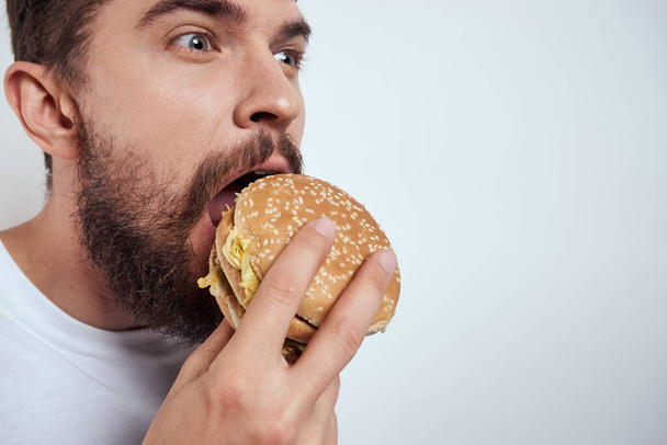 A man eating a hamburger on a light background in a white T-shirt cropped view close-up hunger fast food - Fotografie, Obrázek