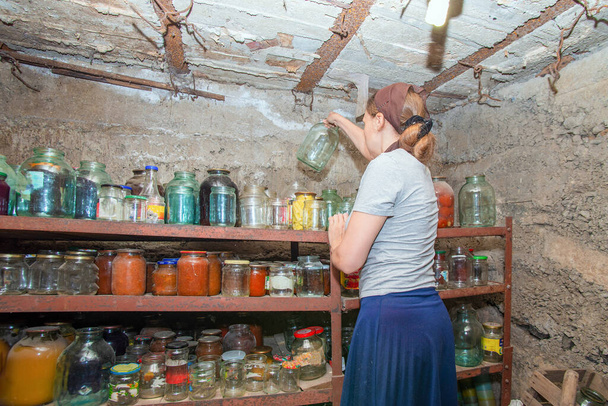 The woman in the cellar is gray-haired, the girl prepares food for the winter, canned food in glass jars on the rack. - Photo, Image
