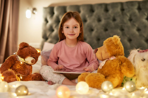 A cute girl wearing pyjamas sitting alone cross-legged on a huge bed barefoot with a tablet gently smiling looking into a camera with her fluffy toys and garlands all over the bed - Foto, Bild