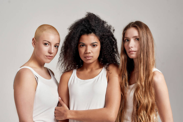 Portrait of three young diverse women wearing white shirts looking at camera while posing together isolated over grey background - Fotografie, Obrázek