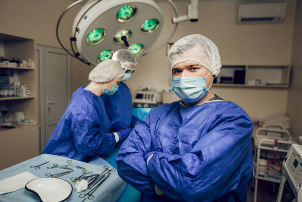 手術室の保護マスクの外科医の肖像画。彼の後ろで他の2人の医者が手術の準備をしている - 写真・画像