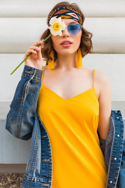 beautiful woman in yellow stylish dress wearing denim jacket, posing with flower, trendy outfit, hippie indie style, spring summer fashion trend, blue sunglasses, street fashion - Фото, зображення