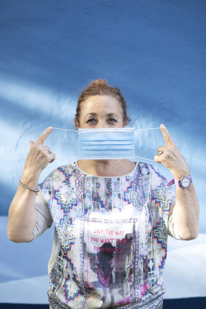 Yaşlı kadın yüzüne tıbbi maske takıyor. Mavi bir duvarın yanında duruyor.. - Fotoğraf, Görsel