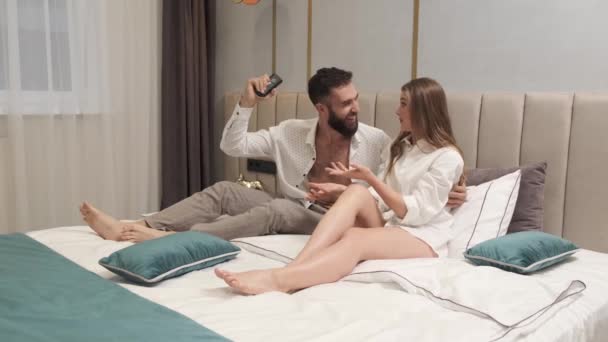 Happy emotional relaxed young family couple lying in bed holding remote control watching tv together. Smiling millennial man and woman talking enjoying television in bedroom at home. - Footage, Video
