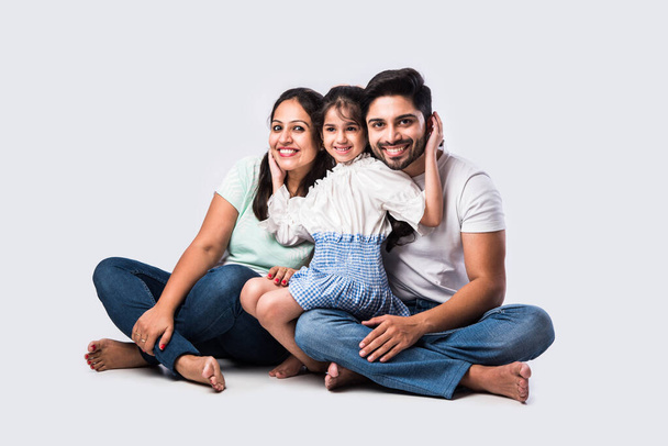 Portrait of Indian Asian young family of four sitting on white flour against white background, looking at camera - Fotó, kép