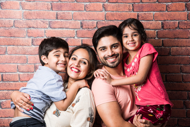 Belle jeune famille indienne de quatre câlins, regardant à la caméra et souriant tout en se tenant contre le mur de briques rouges - Photo, image