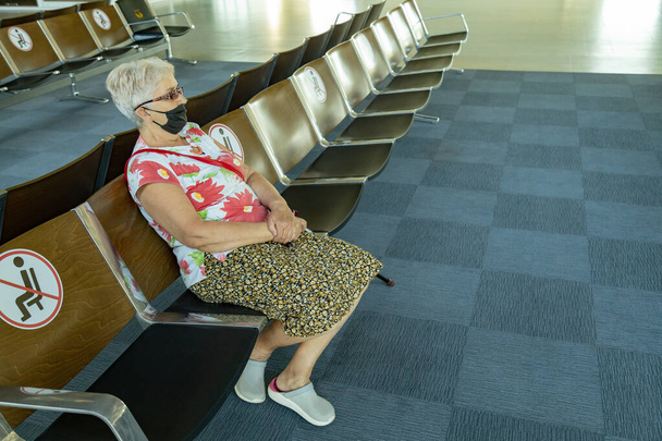 Mulher sênior sentada na área de espera do aeroporto usando máscara facial artesanal. Restrições de viagem devido ao conceito de surto de coronavírus - Foto, Imagem