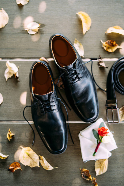 Primer plano de los zapatos negros de un hombre con cordones desatados, sobre una textura de madera con un cinturón y los novios boutonniere, con hojas amarillas de otoño. - Foto, imagen