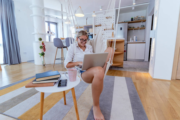 Mooie jonge zakenvrouw die thuis aan een laptop werkt, zittend in een hangstoel in een modern appartement. Lifestyle, modern interieur en werk vanuit huis concept. - Foto, afbeelding