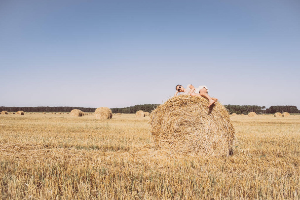 Photo shoot on field, blue sky. Beautiful woman lying on haystack. Summer dream. - 写真・画像