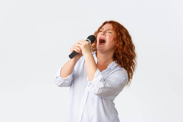 Érzelmek, életmód és szabadidő koncepció. Szenvedélyes és gondtalan vörös hajú női előadó, középkorú nő énekel dalt mikrofonban, énekes játszik karaoke játék, fehér háttér - Fotó, kép