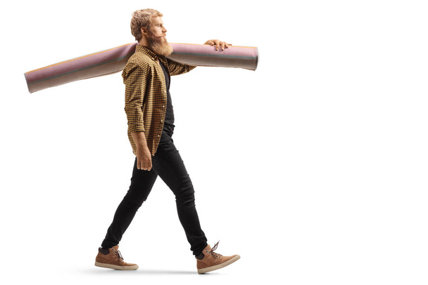 Full length profile shot of a guy walking and carrying a carpet on his shoulder isolated on white background - Foto, Imagen