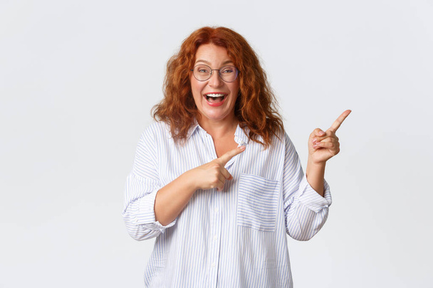 Portrait d'agréable femme souriante d'âge moyen aux cheveux roux, portant des lunettes et un chemisier montrant la publicité, client de l'entreprise recommander un produit ou un service, pointant à droite - Photo, image
