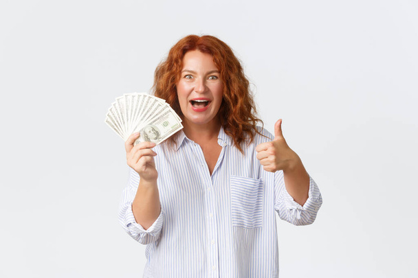 Money, finance and people concept. Excited and amused redhead middle-aged woman showing thumbs-up and smiling thrilled while holding big sum of money, winning cash dollars - Photo, Image