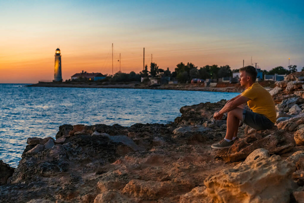  Egy fiatalember ül elgondolkodva egy sziklán, és nézi a naplementét, egy világítótoronnyal a tengerparton a háttérben - Fotó, kép