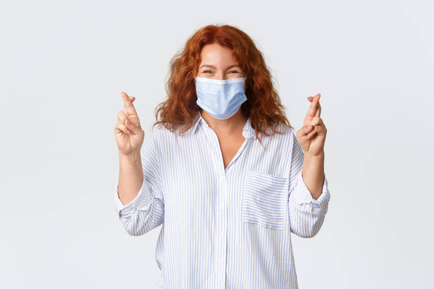 Covid-19 social distancing, coronavirus preventing measures and people concept. Hopeful smiling and optimistic middle-aged woman in medical mask close eyes and cross fingers good luck, making wish - Photo, image