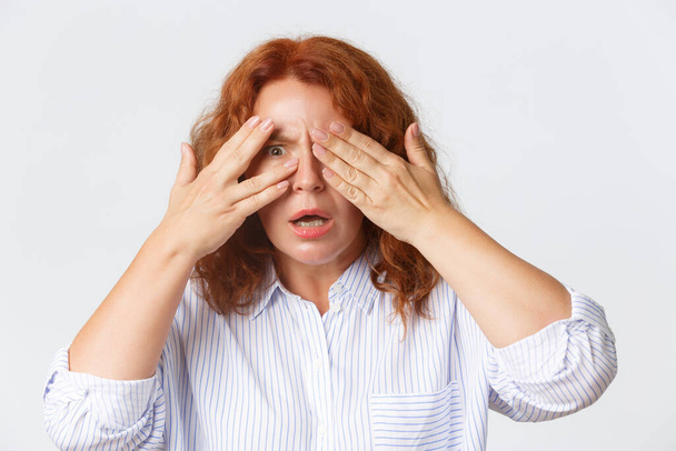 People, emotions and lifestyle concept. Close-up of shocked and alarmed redhead middle-aged woman cover eyes as being scared, but peeking thought fingers curious, white background - Photo, Image