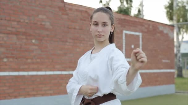 Egy fiatal lány portréja fehér kimonóban, aki karate alapállásba áll egy téglafal hátterében. Lassú mozgás. A kamera egyik oldalról a másikra mozog - Felvétel, videó