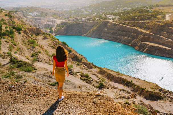  A beautiful woman in red clothes stands over a quarry with an azure lake, quarry in Crimea is a tourist natural attraction - Foto, afbeelding