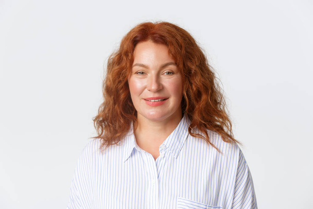 People, emotions and lifestyle concept. Close-up of middle-aged female with red curly hair, wearing casual blouse and smiling at camera cheerful, standing white background - Fotografie, Obrázek