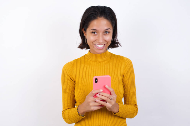 Fénykép gyönyörű fiatal nő használja a mobiltelefon, szörfözik gyors korlátlan internet, kellemes mosoly, piros ingbe öltözött, teszi online vásárlás, elszigetelt sárga - Fotó, kép
