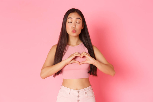 Lifestyle, beauty and women concept. Portrait of romantic beautiful asian girl daydreaming, fell in love, making heart gesture over chest, close eyes and pouting for kiss, standing pink background - Fotografie, Obrázek
