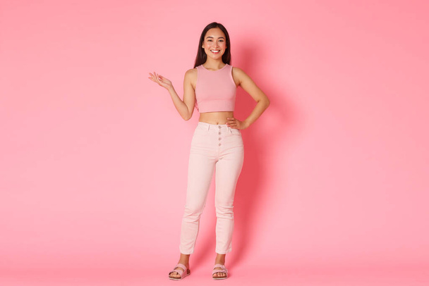 Мода, краса і стиль життя. Портрет привабливої високої азіатської дівчини, яка посміхається і тримає ліву руку підняту, демонструючи продукт на рожевому фоні
 - Фото, зображення