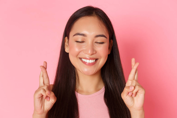 Beauty, fashion and lifestyle concept. Close-up of hopeful smiling, beautiful asian girl close eyes and cross fingers good luck while making wish, praying. Woman having faith in dreams come true - Photo, Image