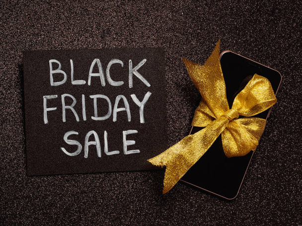 черная меловая доска с надписью "распродажа в черную пятницу" на черном блеске и телефоне с золотой лентой - Фото, изображение
